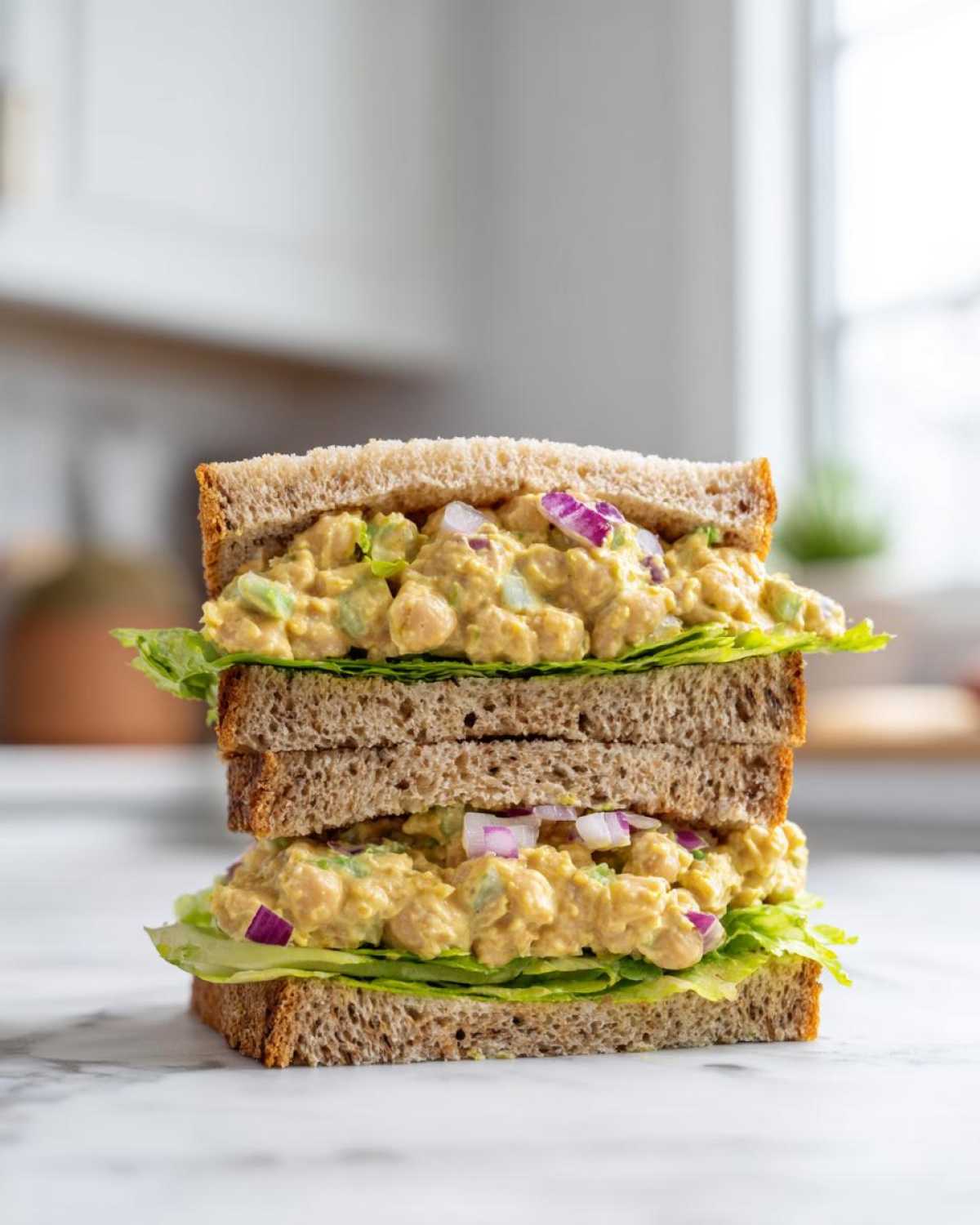 A tall, double-decker Vegan Chickpea Salad Sandwich stacked high on whole wheat bread with lettuce.