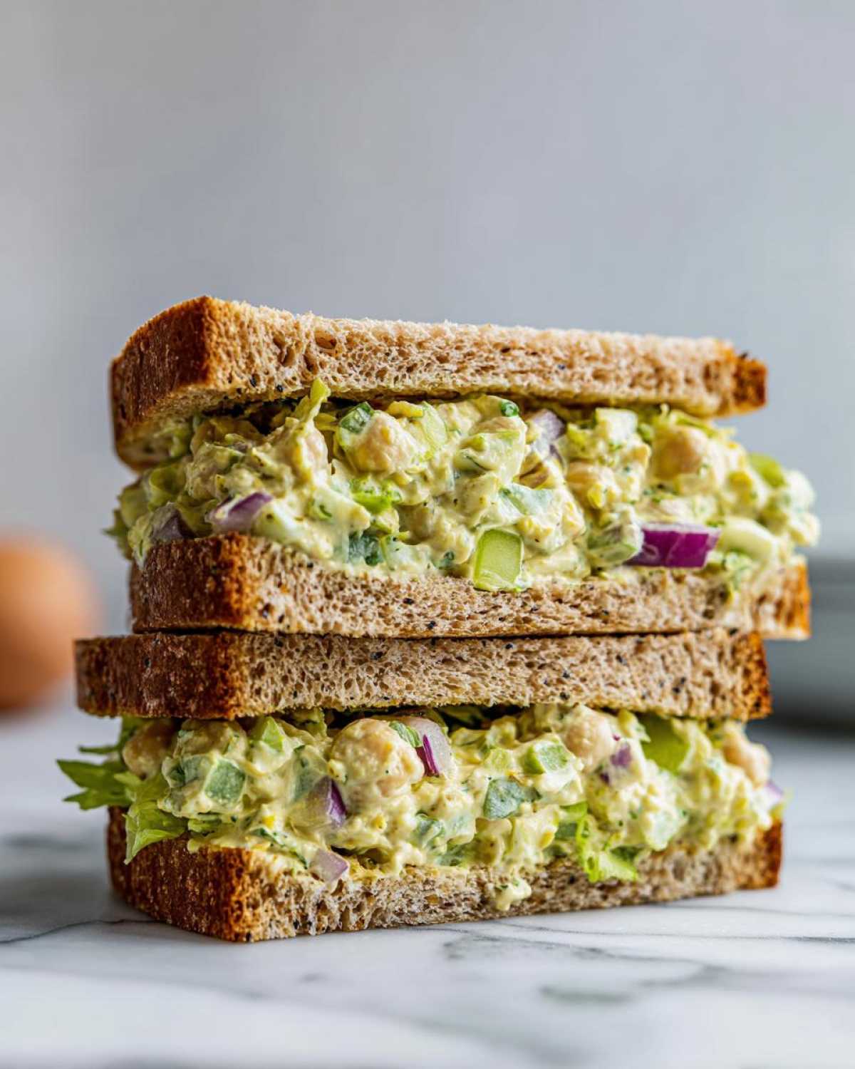 Two halves of a Vegan Chickpea Salad Sandwich stacked, showing creamy filling with chickpeas, celery, and red onion on whole wheat bread.
