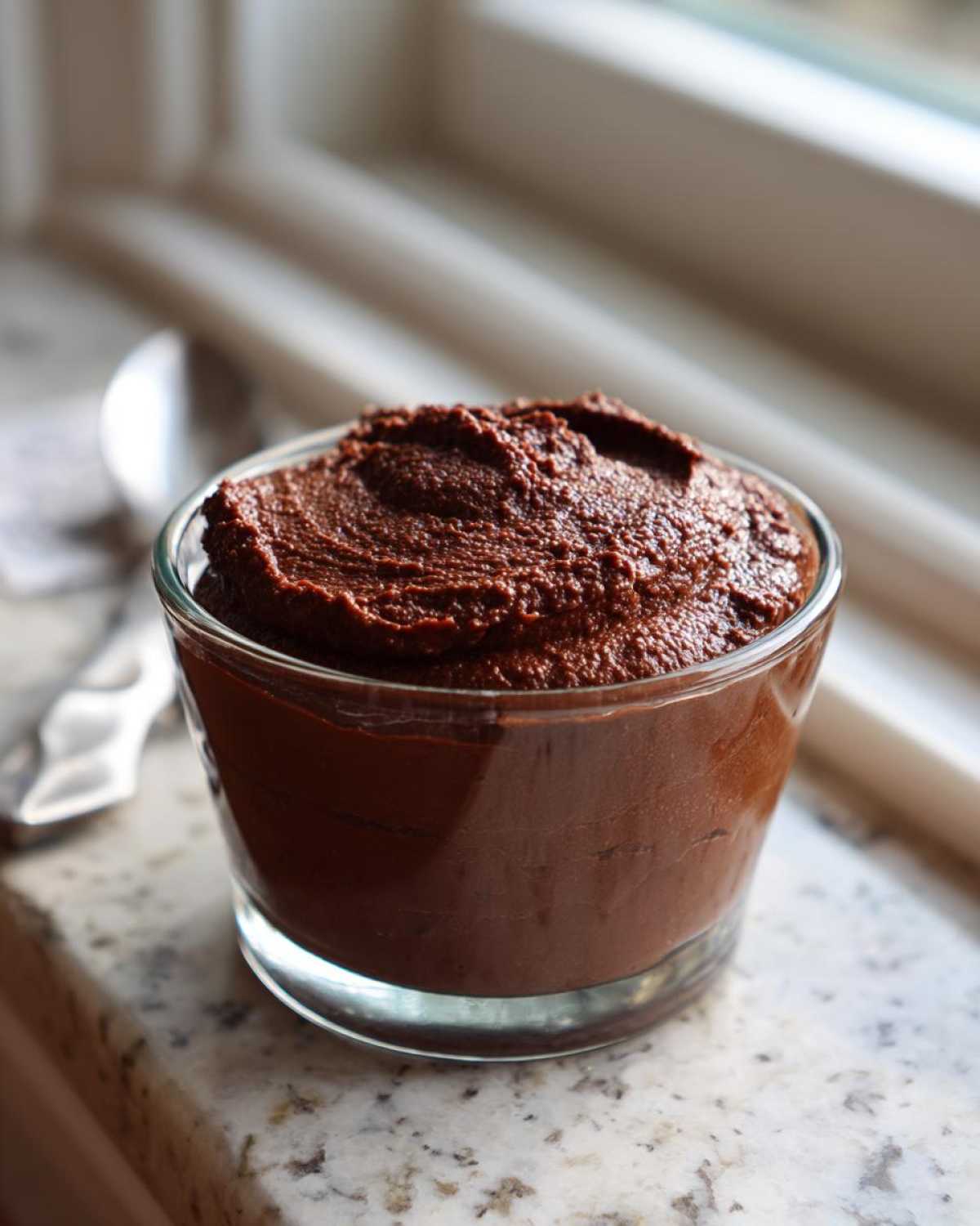 A glass filled with rich, dark Vegan Chocolate Avocado Mousse, sitting on a light countertop.