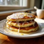 A stack of three golden Vegan Lemon Poppy Seed Pancakes drizzled with a thick lemon glaze and topped with zest.