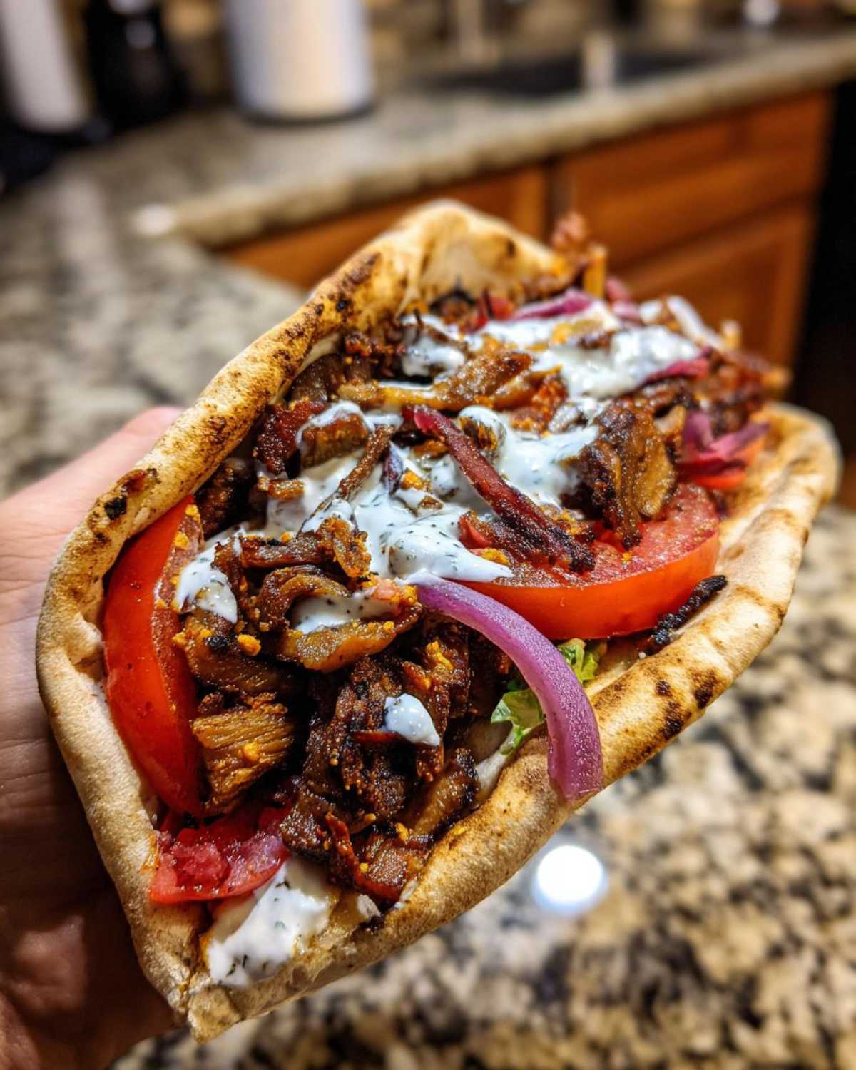 A hand holding a generously filled Vegan Mushroom Shawarma in a toasted pita bread, topped with sauce and onions.