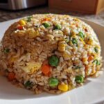 A perfectly molded dome of Vegetable Fried Rice with mixed peas, carrots, corn, and scrambled egg on a white plate.