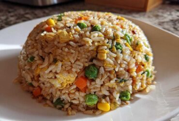 A perfectly molded dome of Vegetable Fried Rice with mixed peas, carrots, corn, and scrambled egg on a white plate.