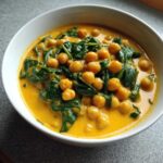 A close-up of a white bowl filled with vibrant Vegetarian Chickpea Curry with Coconut Milk, featuring chickpeas and wilted spinach.