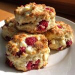 Close-up of four flaky White Chocolate Raspberry Scones stacked on a white plate, showing fresh raspberries and white chocolate chips.