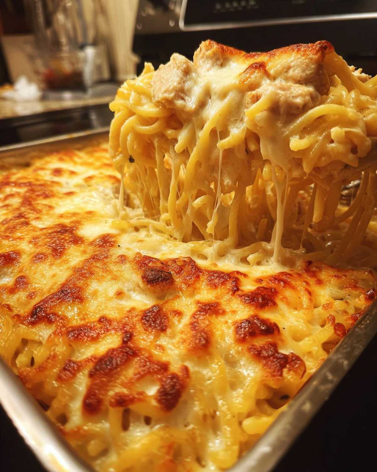 A serving spoon lifts a portion of Cheesy Chicken Alfredo Bake, showing long, gooey cheese strings stretching from the golden-brown baked dish.
