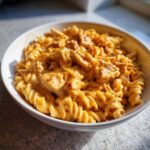 A close-up of a white bowl filled with creamy Cheesy Chicken Queso Pasta made with rotini noodles and chunks of chicken.