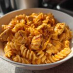 A close-up of a white bowl filled with creamy Cheesy Chicken Queso Pasta using rotini noodles and chunks of seasoned chicken.