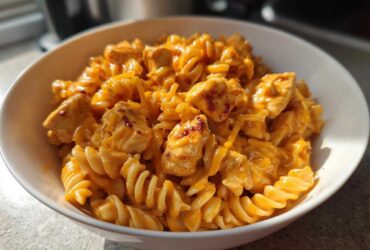 A close-up of a white bowl filled with creamy Cheesy Chicken Queso Pasta using rotini noodles and chunks of seasoned chicken.