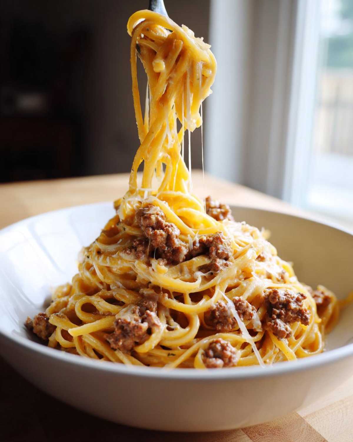 A fork lifts spaghetti coated in garlic cream sauce and ground beef from a bowl of Cheesy Ground Beef Pasta, showing cheese pull.