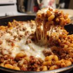 A fork lifts a portion of Cheesy Ground Beef Pasta Skillet with Mozzarella, showing dramatic cheese pulls over the skillet.