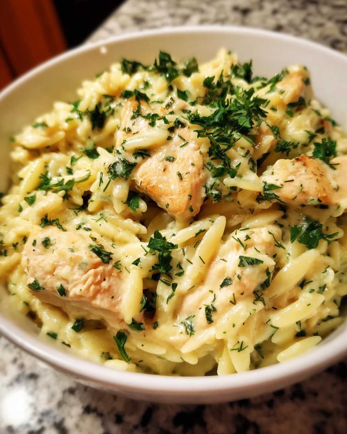 A close-up of a white bowl filled with Creamy Lemon Chicken Orzo, topped generously with fresh chopped parsley.
