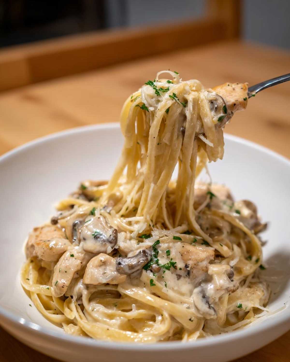 A fork lifting a generous portion of Creamy Mushroom Chicken Alfredo Pasta from a white bowl, showing fettuccine coated in sauce.