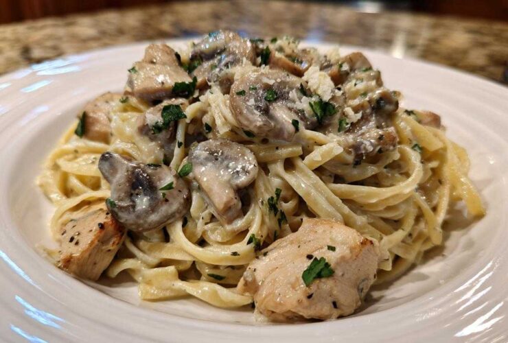 A close-up of a white plate piled high with Creamy Mushroom Chicken Alfredo Pasta, topped with mushrooms and parsley.