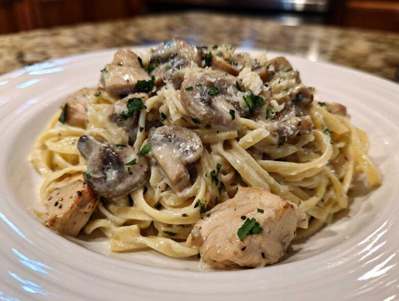 A close-up of a white plate piled high with Creamy Mushroom Chicken Alfredo Pasta, topped with mushrooms and parsley.