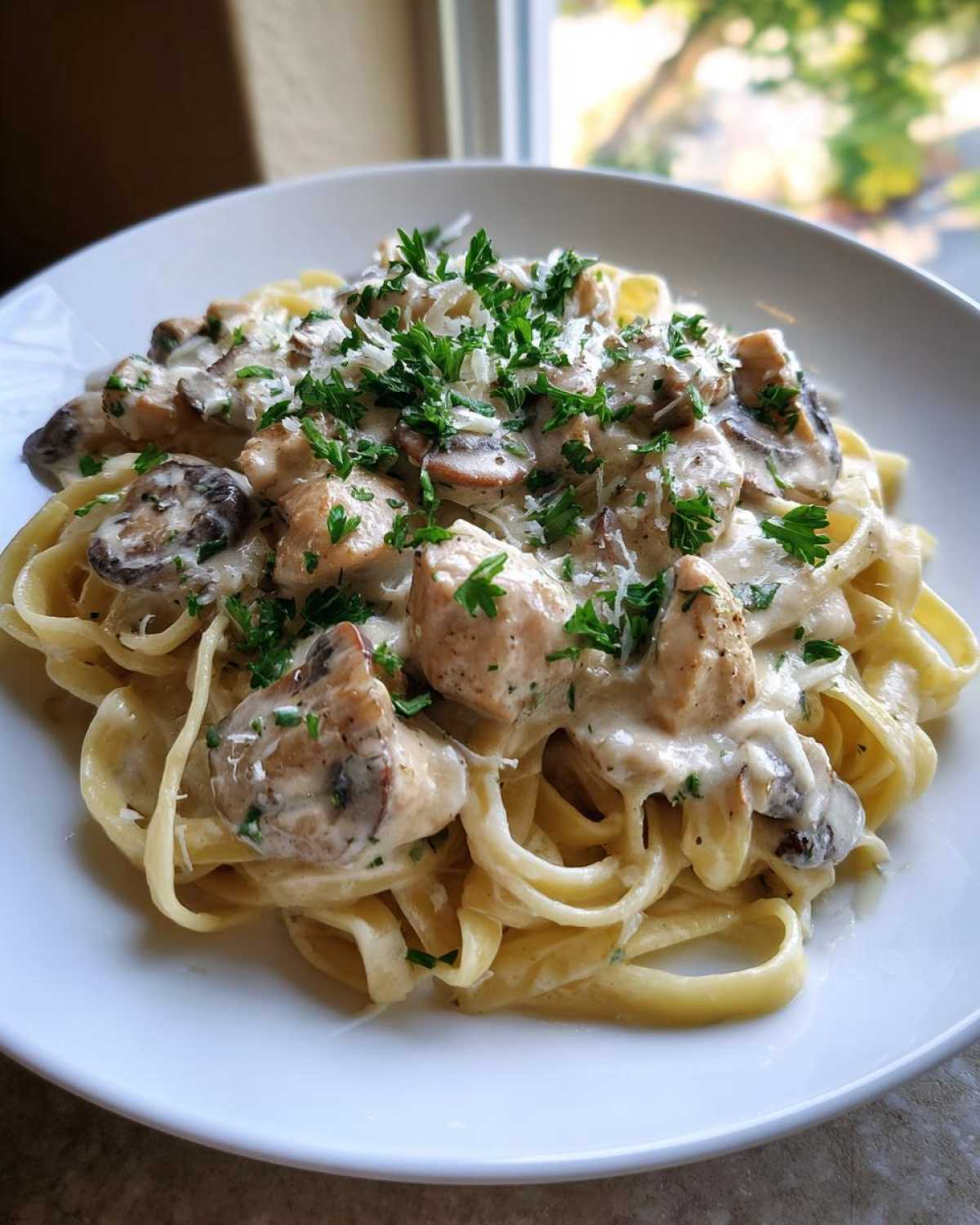 A close-up of a white bowl filled with Creamy Mushroom Chicken Pasta with Parmesan, topped with fresh parsley.