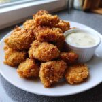 A plate piled high with golden brown Crispy Chicken Nuggets next to a small white ramekin of creamy garlic dip.