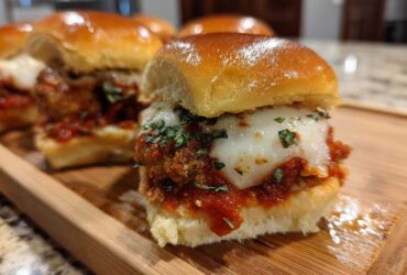 A close-up of one of the Crispy Chicken Parmesan Sliders with melted mozzarella cheese and parsley on a wooden board.
