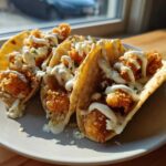 Three crispy chicken tacos drizzled with garlic lime sauce, served on a light gray plate near a window.