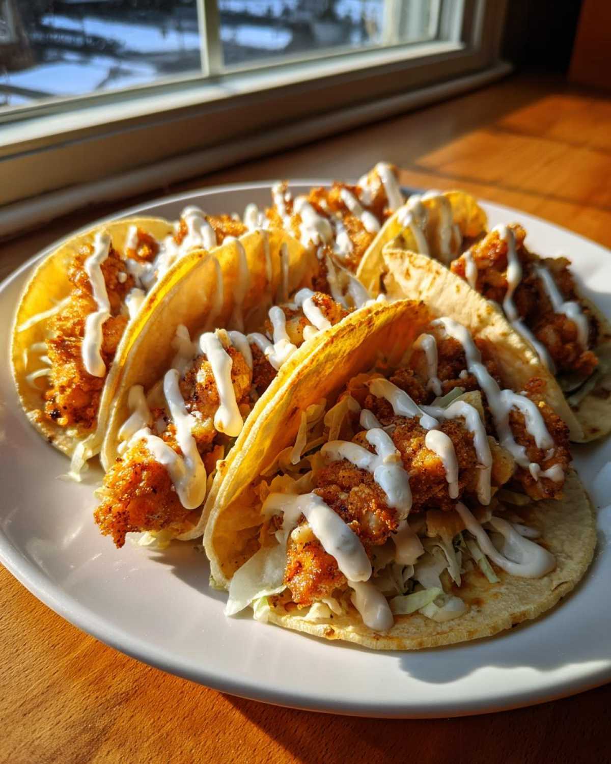 A plate of several Crispy Chicken Tacos filled with shredded lettuce and drizzled generously with a white garlic lime sauce.