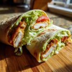 A halved Crispy Chicken Wrap with Avocado Sauce showing crispy chicken, lettuce, and tomato inside a toasted tortilla.