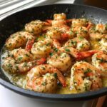 Close-up of plump shrimp cooking in a skillet with rich garlic butter and lemon sauce, garnished with parsley.
