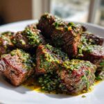Close-up of juicy Garlic Butter Steak Bites generously topped with a bright green herb sauce on a white plate.
