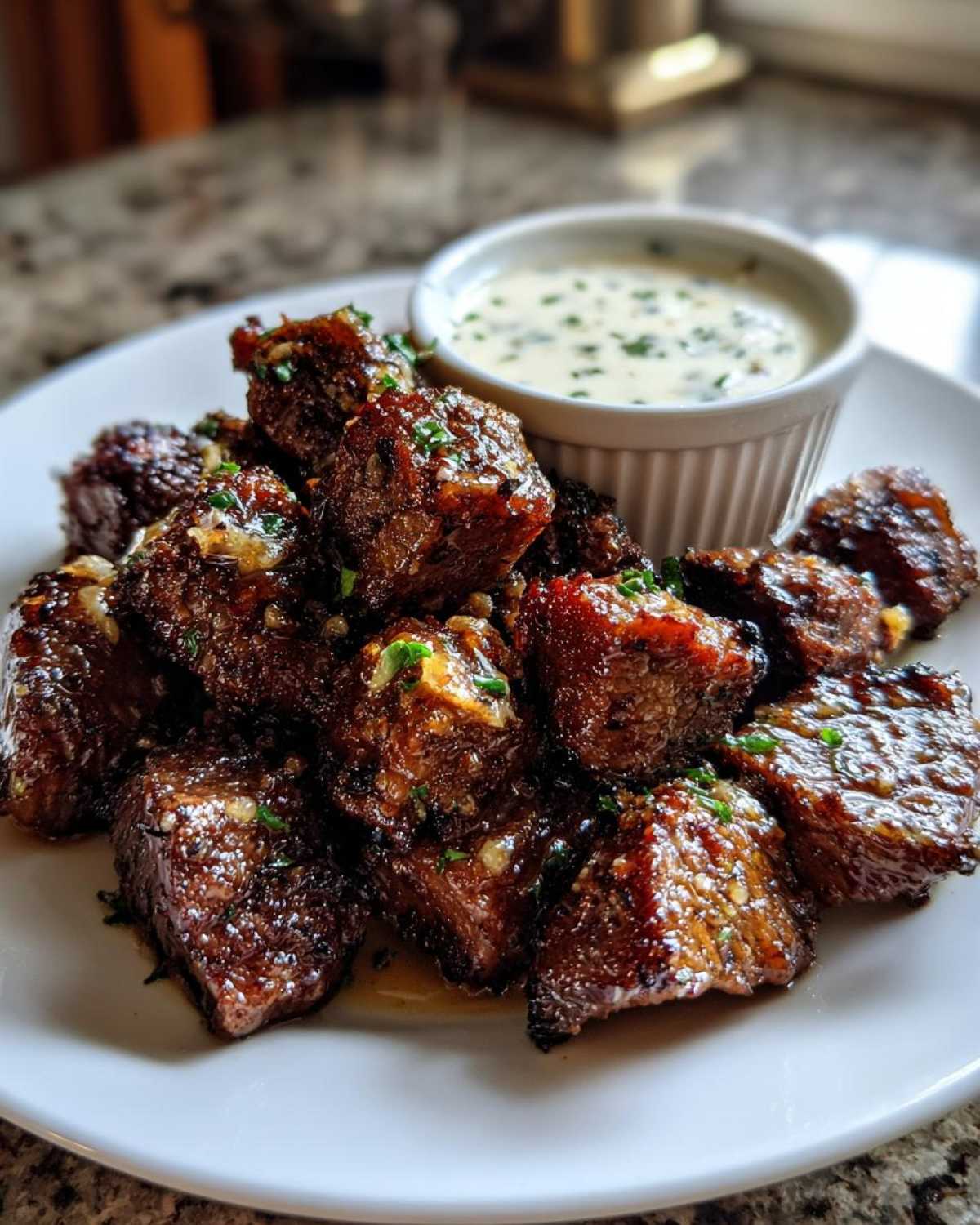 A plate piled high with glistening Garlic Butter Steak Bites, garnished with herbs, next to a small bowl of creamy garlic sauce.
