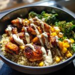 A close-up of a Honey Mustard Chicken Bowl featuring glazed chicken pieces over quinoa, topped with corn, broccoli, and a creamy sauce drizzle.