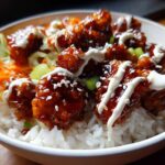 Close-up of a Honey Soy Chicken Bowl featuring glazed chicken pieces over white rice, drizzled with white sauce and sesame seeds.