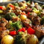 Close-up of browned Garlic Butter Chicken Bites mixed with roasted broccoli, red peppers, and small potatoes on a sheet pan.