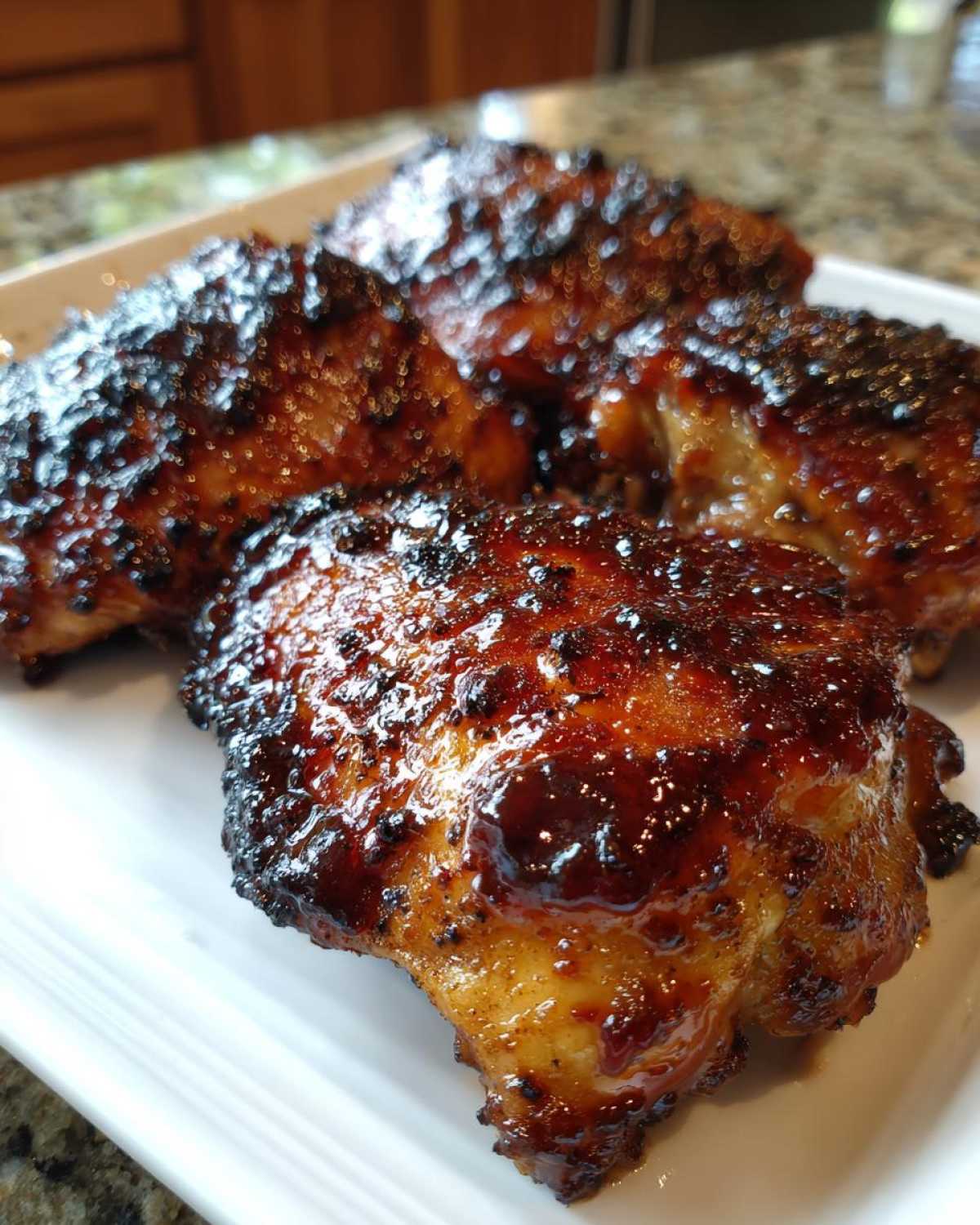 Four pieces of Sticky BBQ Chicken Thighs with Crispy Edges, glazed with a thick, dark sauce, served on a white platter.