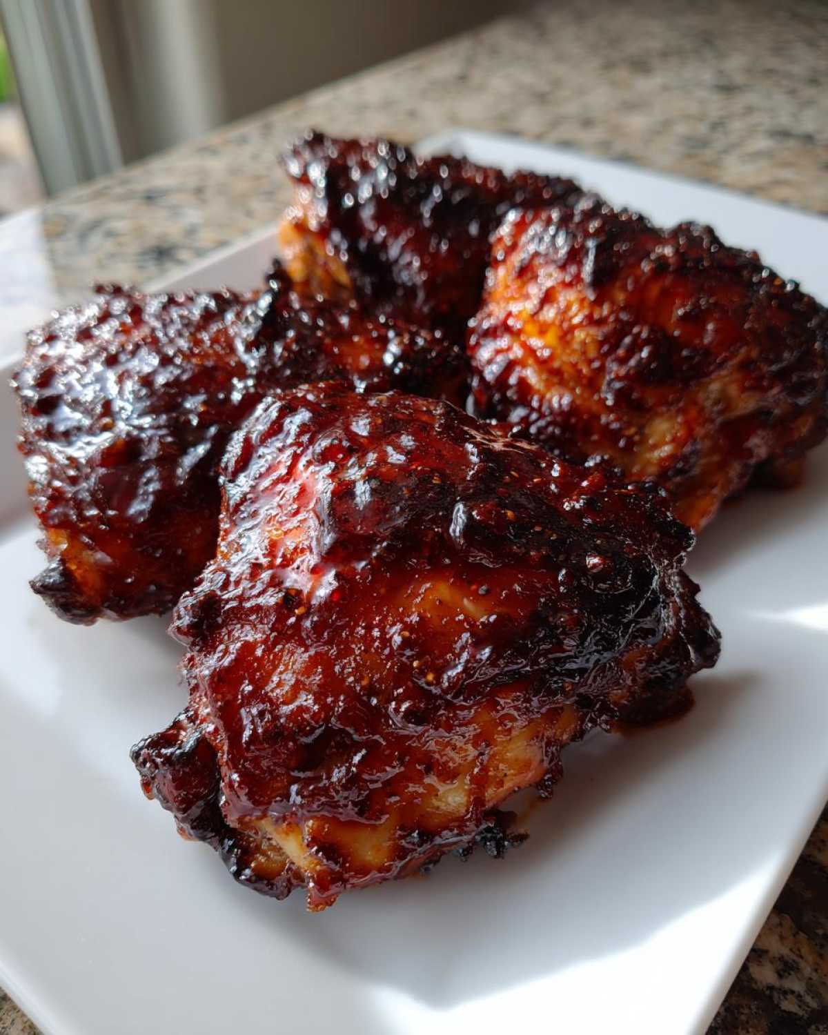 Three pieces of Sticky BBQ Chicken Thighs with Crispy Edges, coated in a thick, dark, glossy sauce, served on a white plate.