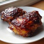 Two perfectly cooked Sticky BBQ Chicken Thighs glistening with a dark, thick honey glaze on a white plate.