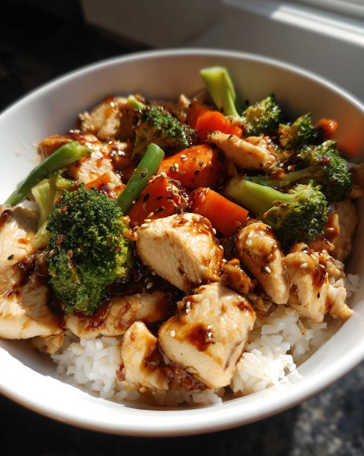 A close-up of a white bowl filled with Sticky Teriyaki Chicken Bowl with Rice and Veggies, topped with sesame seeds.