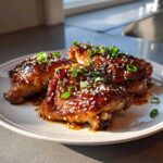 Three pieces of Sticky Teriyaki Chicken Thighs with Garlic, coated in a thick glaze and topped with sesame seeds and green onions.