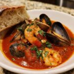 A bowl of vibrant red tomato-based Cioppino stew featuring shrimp, mussels, clams, and a side of crusty bread.