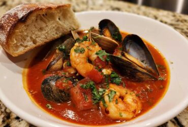 A bowl of vibrant red tomato-based Cioppino stew featuring shrimp, mussels, clams, and a side of crusty bread.