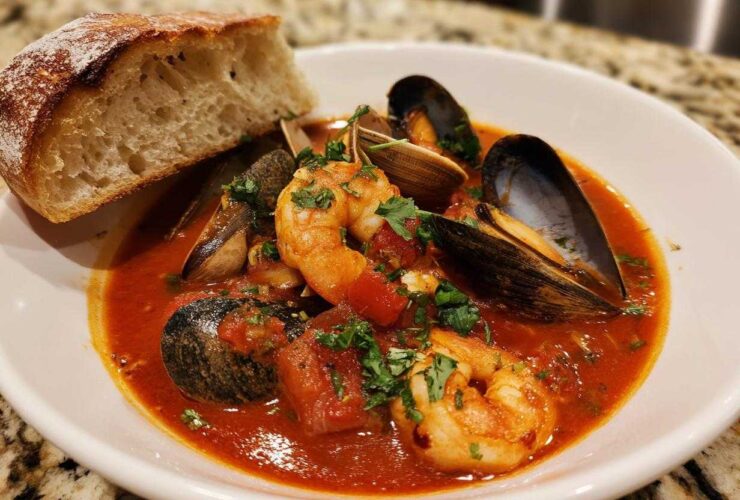 A bowl of vibrant red tomato-based Cioppino stew featuring shrimp, mussels, clams, and a side of crusty bread.