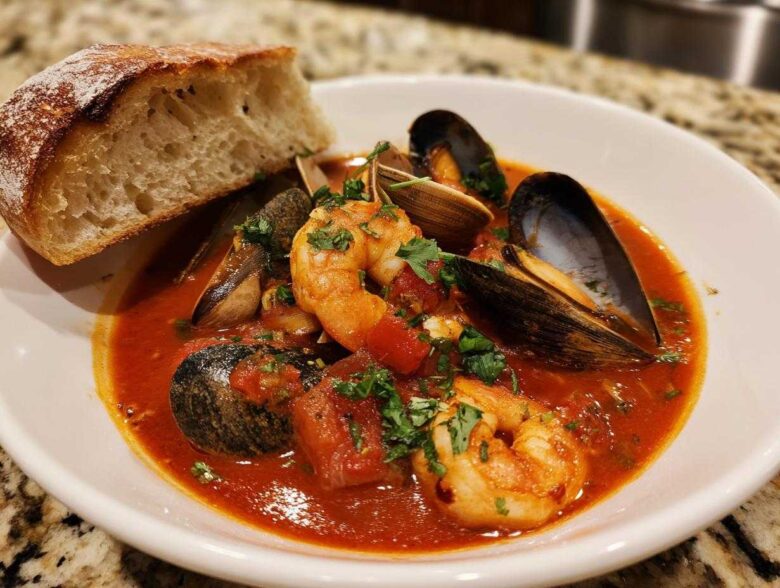 A bowl of vibrant red tomato-based Cioppino stew featuring shrimp, mussels, clams, and a side of crusty bread.
