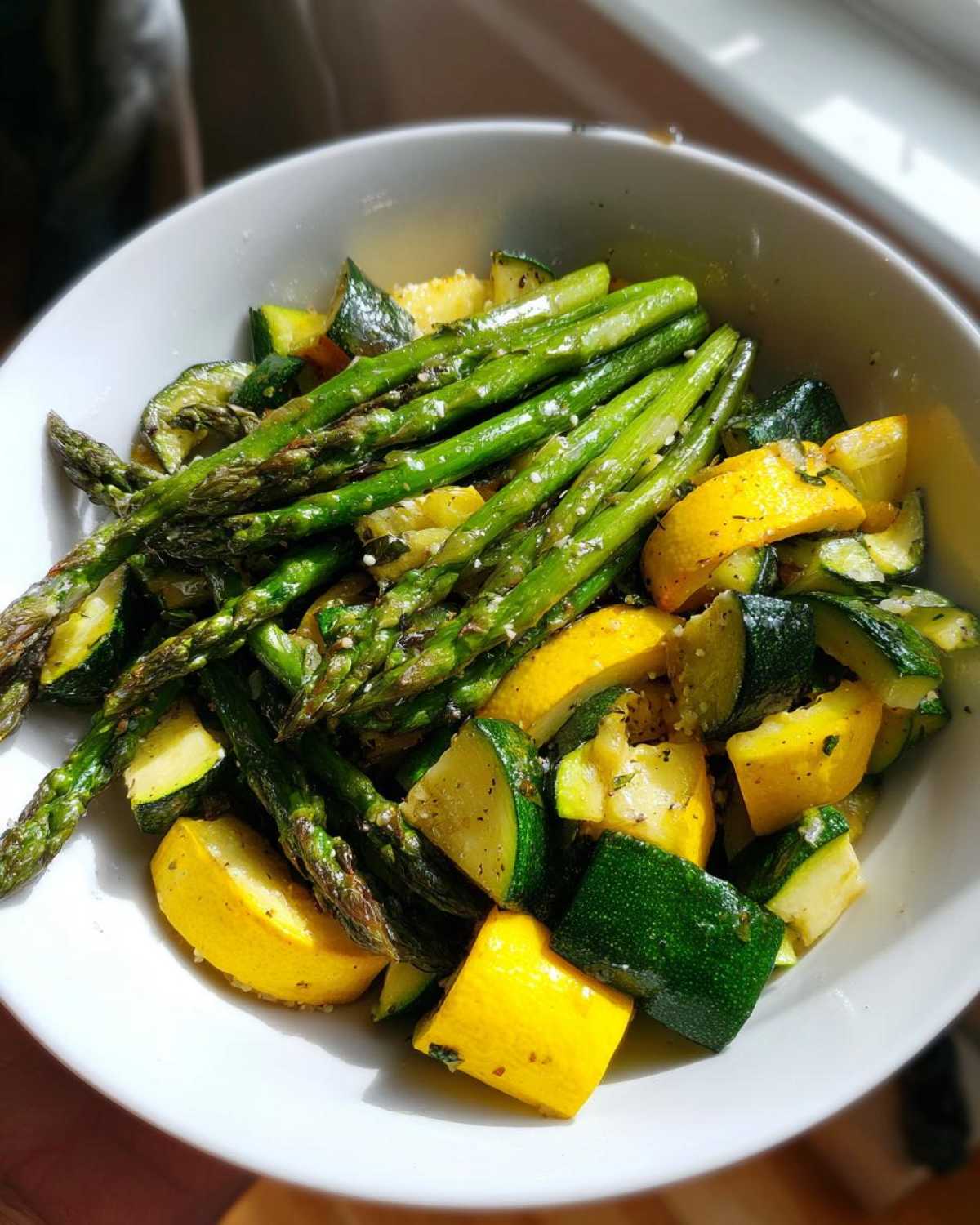 A white bowl filled with bright green asparagus spears mixed with chopped yellow squash and zucchini, seasoned lightly.