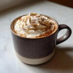 A cozy, dark mug filled with a Banana Bread Latte, generously topped with whipped cream and a sprinkle of cinnamon.