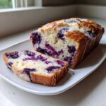 A slice cut from a moist Blueberry Cream Cheese Bread Loaf, showing rich purple blueberries throughout the crumb and a light glaze.