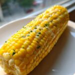 A close-up of a glistening ear of Buttered Corn on the cob, topped with melted butter and chopped parsley.
