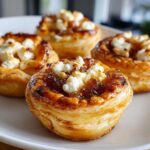 Close-up of four flaky Caramelized Onion and Goat Cheese Tarts topped with crumbled goat cheese on a white plate.