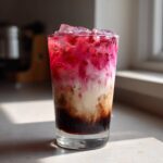 A tall glass showing distinct layers of a Cherry Vanilla Iced Latte with dark syrup, creamy milk, and bright pink cherry topping over ice.
