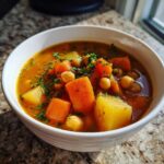 A close-up of a hearty bowl of Chickpea Potato Soup, featuring large chunks of carrots, potatoes, and chickpeas in a rich broth, topped with parsley.
