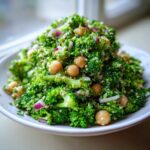 A close-up of a vibrant Chopped Broccoli and Chickpea Salad piled high on a white plate, featuring bright green broccoli florets, chickpeas, and red onion.