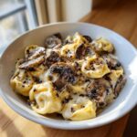 A white bowl filled with rich Creamy Mushroom Tortellini Alfredo, topped with sliced sautéed mushrooms and cracked pepper.