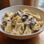 A close-up of a white bowl filled with Creamy Mushroom Tortellini Alfredo, featuring tortellini coated in rich sauce and topped with sliced mushrooms.